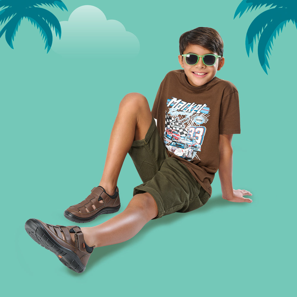 Niño con sandalias cerradas color café, camiseta gráfica y lentes de sol en fondo veraniego.