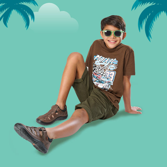 Niño con sandalias cerradas color café, camiseta gráfica y lentes de sol en fondo veraniego.