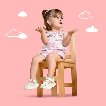 Bebé niña sentada en silla de madera con sandalias blancas coqueta y audaz, y con conjunto rosa a cuadros.