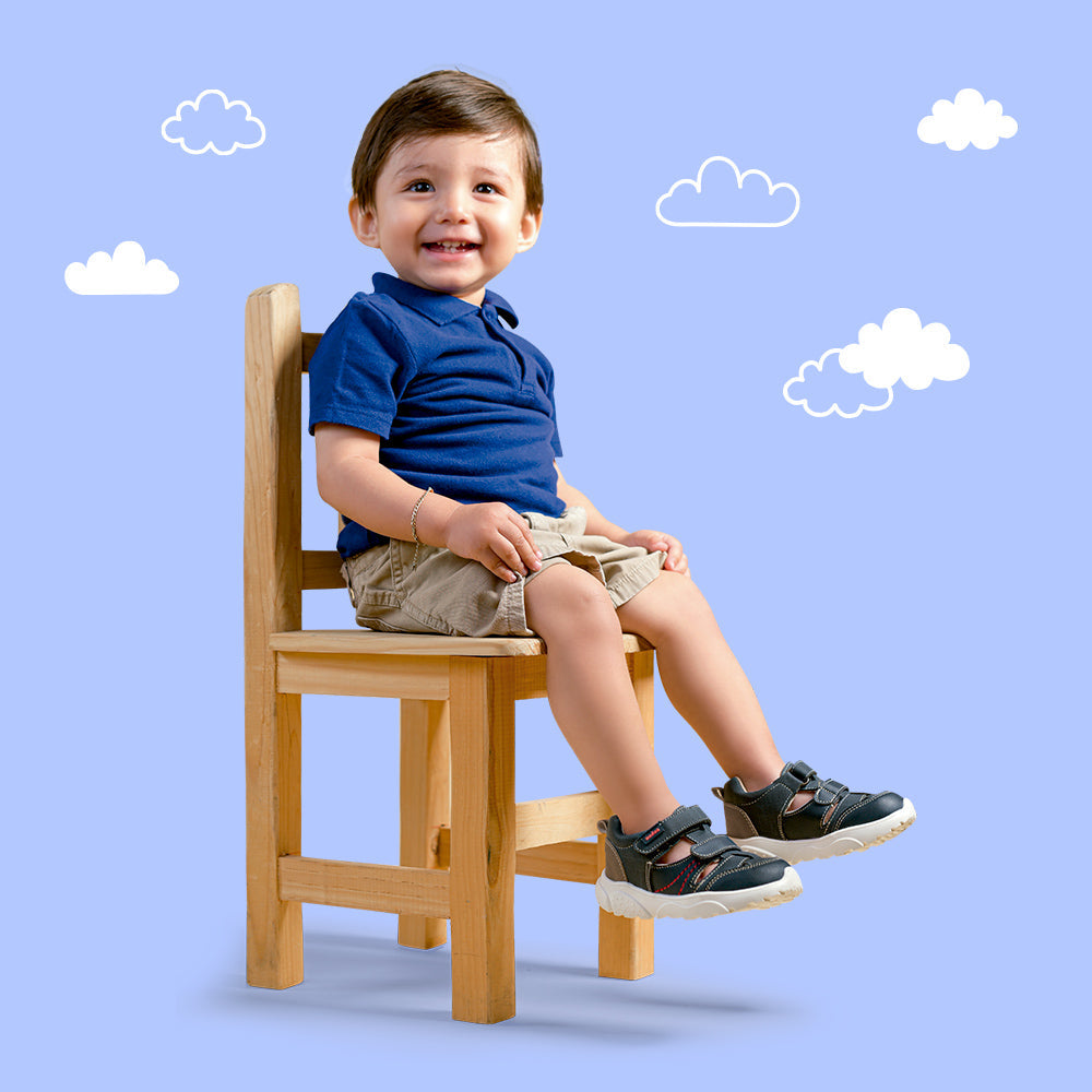 Bebé niño sonriendo sentado en silla de madera, con polo azul, shorts beige y tenis deportivos oscuros coqueta y audaz.